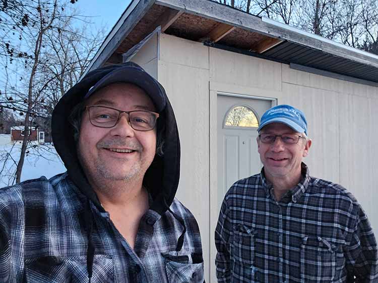 Pete and Roger, the two founders of Minnesota Mobile Sauna, wearing warm outdoor clothing and standing outside in front of an off-white mobile sauna unit during a winter day in Minnesota.