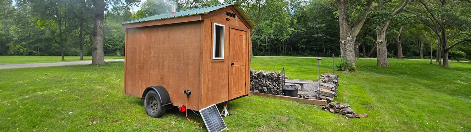 A panoramic wide-angle view of a wood-sided mobile sauna parked on a lush green lawn. A portable solar panel is propped up in the foreground, and a neatly stacked woodpile with a bench is visible in the background under a canopy of trees.