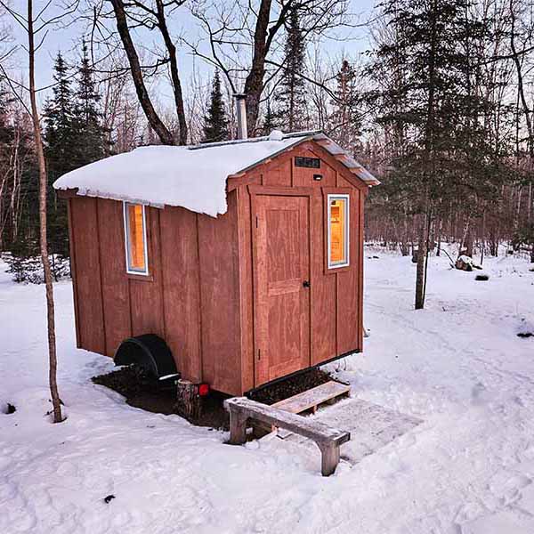 An exterior shot of a mobile wood-fired sauna on a trailer, parked in a deep snowy wooded clearing at dusk. The warm glow of the interior lights shines from two vertical windows, contrasting sharply with the snow on the roof and ground. A small wooden step bench is set up in the foreground, and the stainless steel chimney stack is visible.