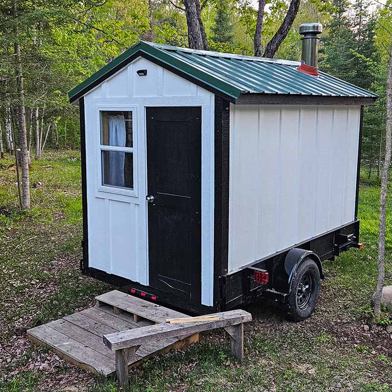 An exterior shot of a mobile wood-fired sauna on a trailer, featuring distinctive white board and batten siding with black trim and a green metal roof. A rustic wooden entry step is set up in front of the door in a natural, wooded clearing.