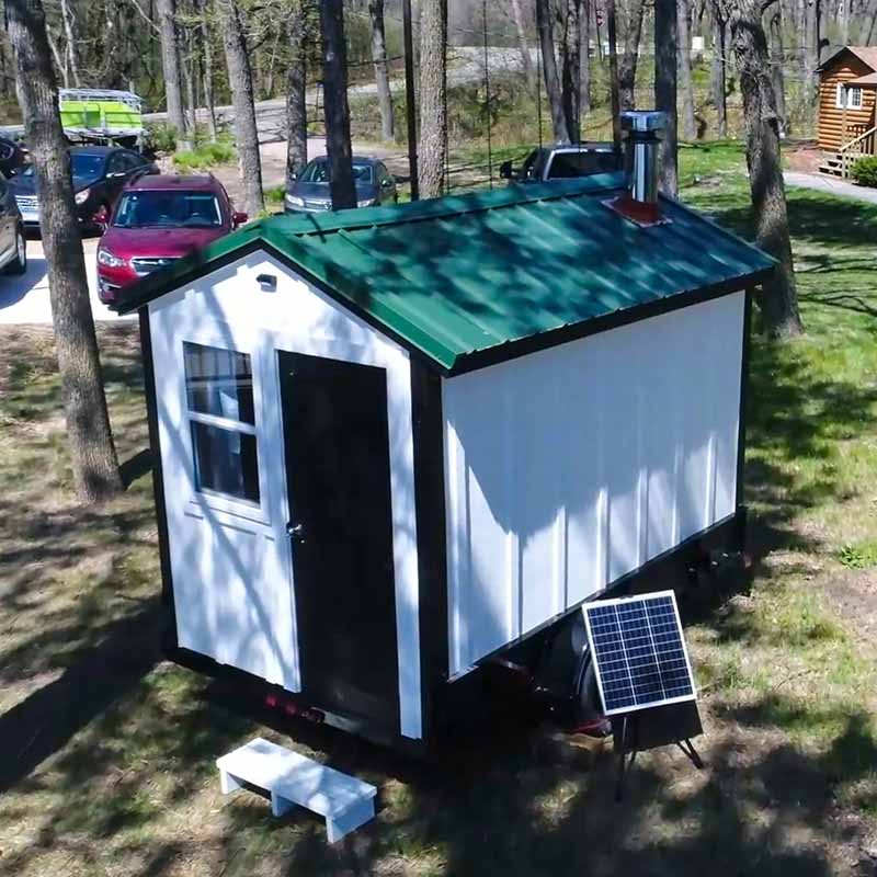 A drone's-eye view of a white mobile sauna on a trailer, featuring a distinctive green metal roof and an outdoor chimney stack. The portable solar panel is visible, showcasing the unit's off-grid capabilities in a shaded, grassy location next to a tree line.