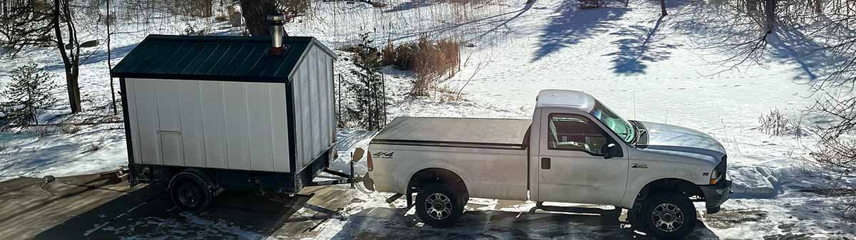 A side profile view of a white pickup truck and a mobile wood-fired sauna. The truck and trailer are parked in a snowy residential driveway in Minnesota, demonstrating the logistics of winter delivery. The stainless steel chimney stack is visible on the sauna.