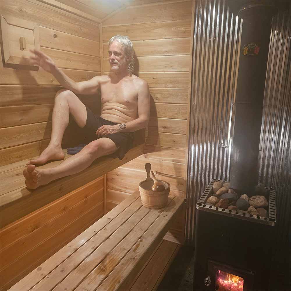 An interior view of a cedar-lined mobile sauna with a man relaxing on the elevated top bench. A wood-burning stove with hot rocks is visible, along with a wooden water bucket and ladle, and the adjustable ventilation hatch.