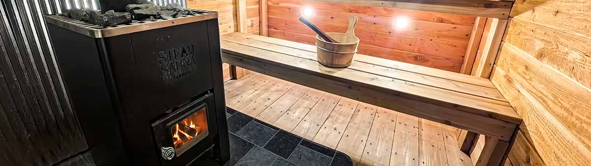 A panoramic wide-angle view of a mobile sauna interior. On the left is a black wood-burning stove with a visible fire in the glass door, topped with sauna rocks. On the right are two-tiered cedar benches with a wooden water bucket and ladle. The floor is also made from cedar.