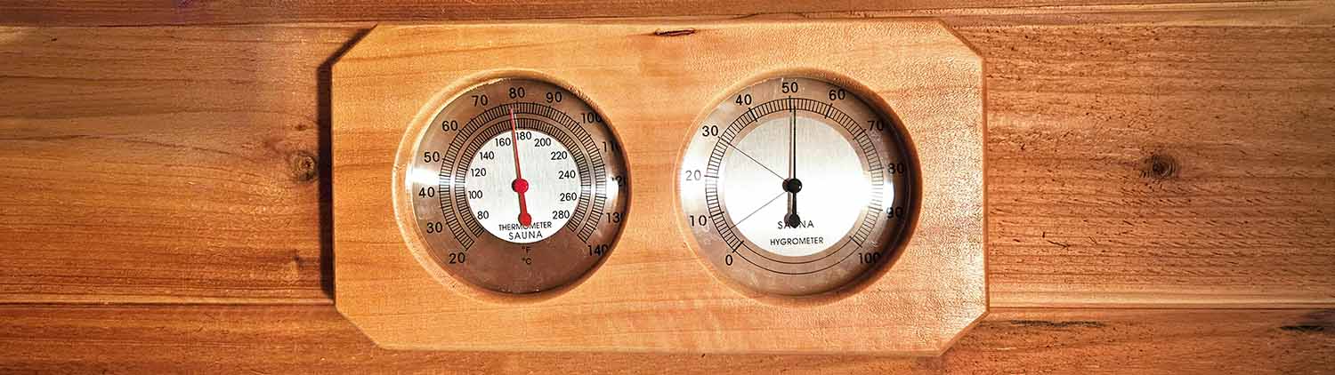 A panoramic close-up of a traditional wooden-mounted sauna thermometer and hygrometer inside a cedar-lined room. The thermometer needle is pointing to 180 degrees Fahrenheit, and the hygrometer shows the humidity level.