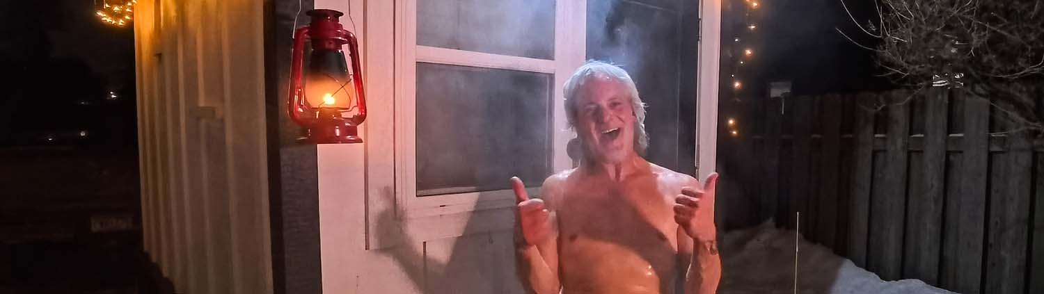 A panoramic wide-angle photograph taken at night. A happy, bare-chested man with grey hair, his skin glistening with moisture and steam, is emerging from the mobile sauna door. He is smiling broadly and giving two enthusiastic thumbs up. A glowing red lantern hangs next to him, and a snowy backyard is visible in the distance.