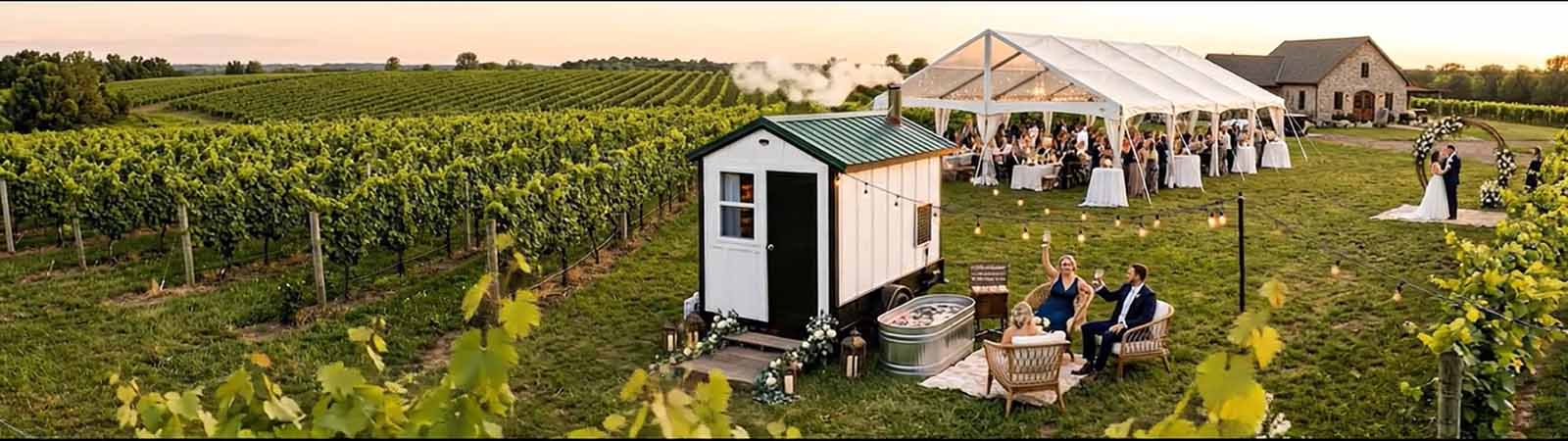 Wide-angle view of a white mobile sauna unit staged at a luxury vineyard wedding with a sailcloth reception tent and vineyard rows in the background.