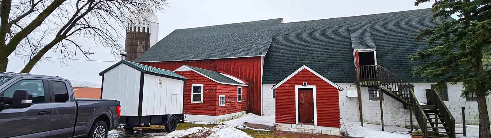 A panoramic, ultra-wide view of a Minnesota Mobile Sauna unit being towed by a grey pickup truck in front of a rustic red barn complex with a silo and snow-covered ground.